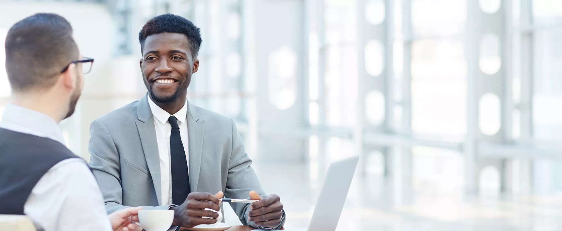 Two men smiling during a meeting. The structured metal window frames in the background illuminate the office brightly. The is a laptop and notebook on the table between them. Two men smiling during a meeting. The structured metal window frames in the background illuminate the office brightly. The is a laptop and notebook on the table between them.