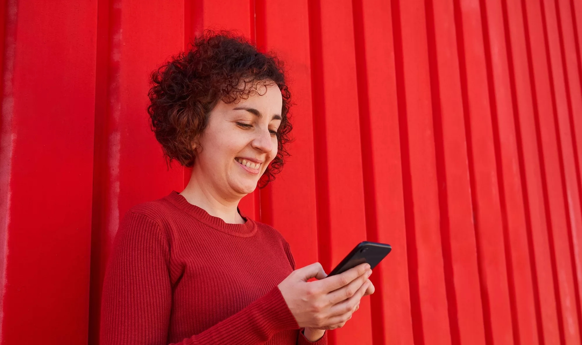 A smiling woman looking down at her phone, leaning against a red, slatted wall. A smiling woman looking down at her phone, leaning against a red, slatted wall.