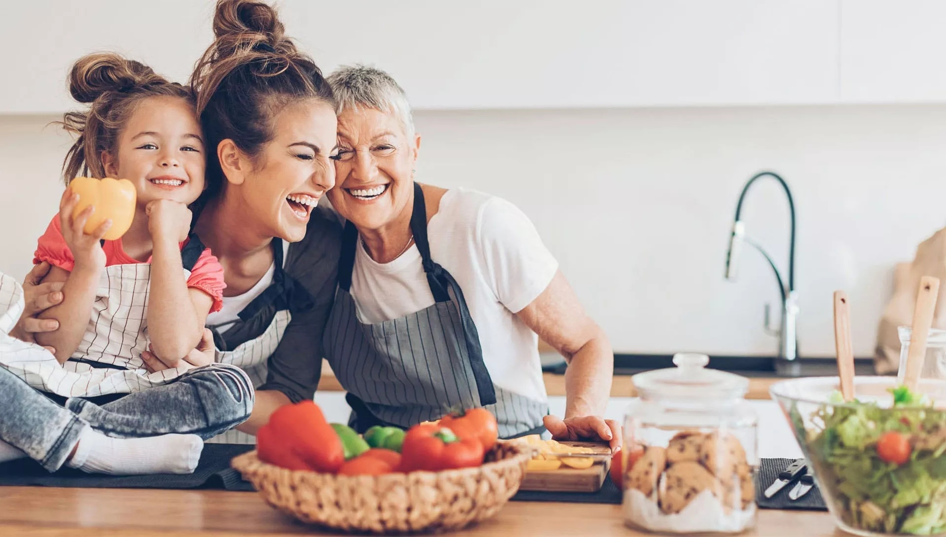 A Grandmother, a mother, and a child smiling in the kitchen, mid-preparation of a meal. A Grandmother, a mother, and a child smiling in the kitchen, mid-preparation of a meal.