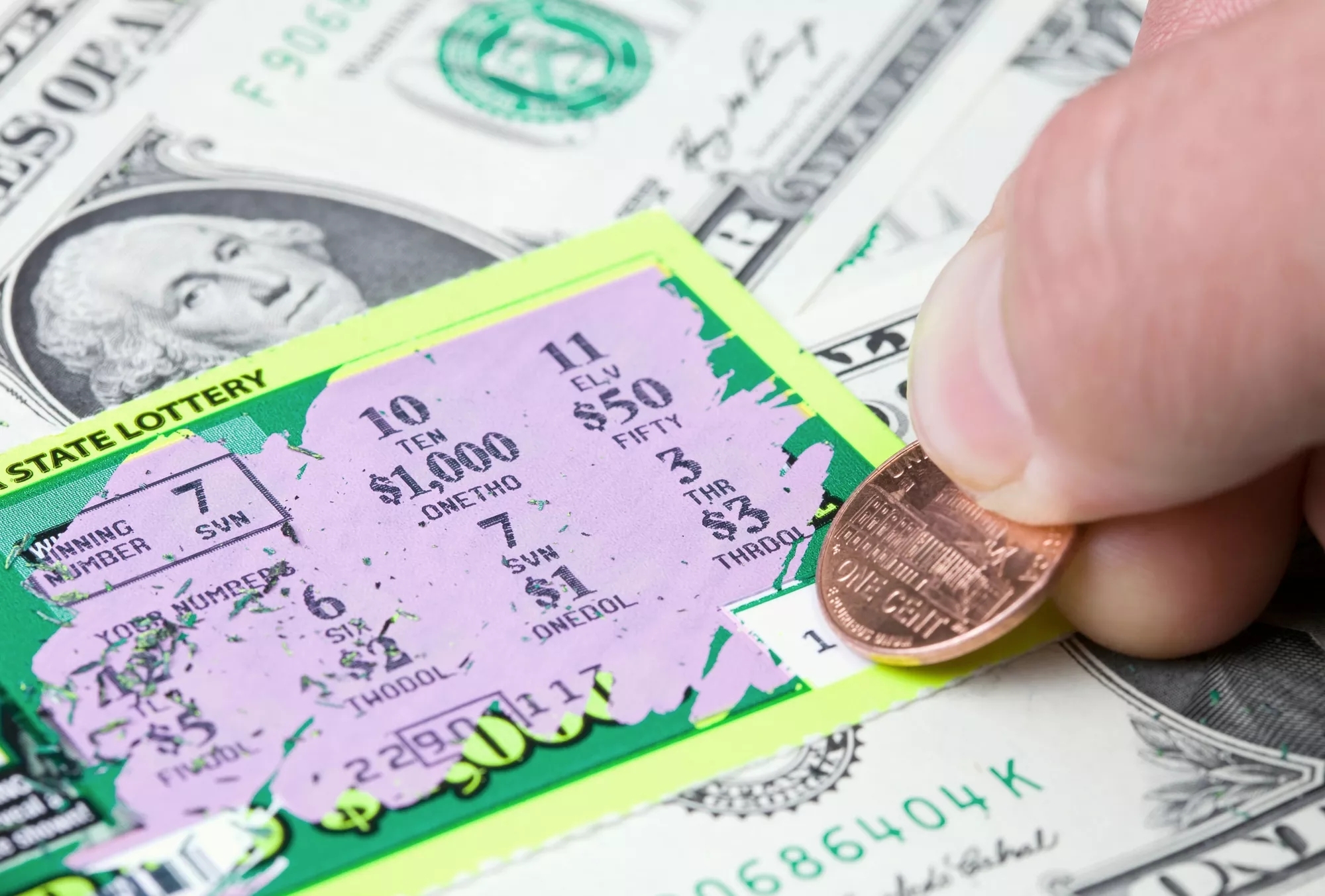 A lottery ticket being scratched with a penny, on top of spread out one dollar bills on the table below. A lottery ticket being scratched with a penny, on top of spread out one dollar bills on the table below.
