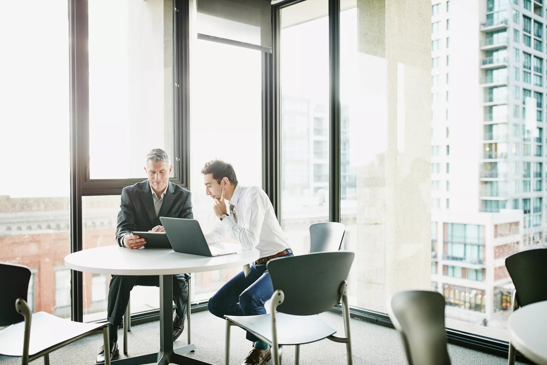 Two men meeting at a small table by an office window looking out on the city. They are discussing information on the laptop and tablet in front of them. Two men meeting at a small table by an office window looking out on the city. They are discussing information on the laptop and tablet in front of them.