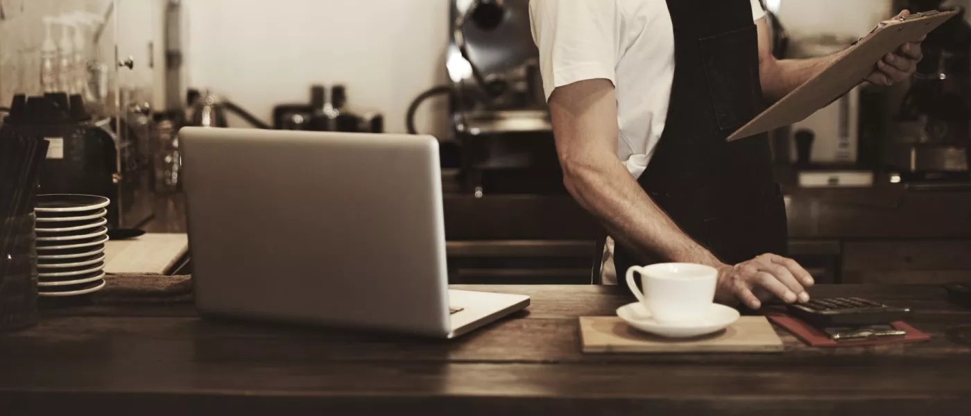 An employee of a coffeeshop working on a laptop, clipboard and calculator behind the counter.