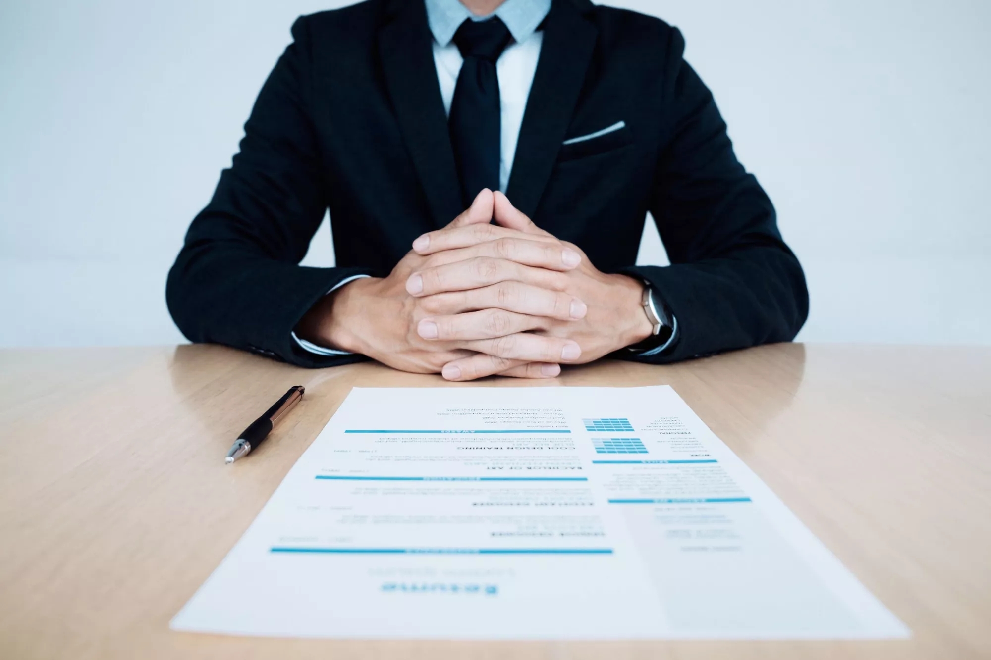 A person wearing a suit sits across the table, an application form and pen in front of them. A person wearing a suit sits across the table, an application form and pen in front of them.