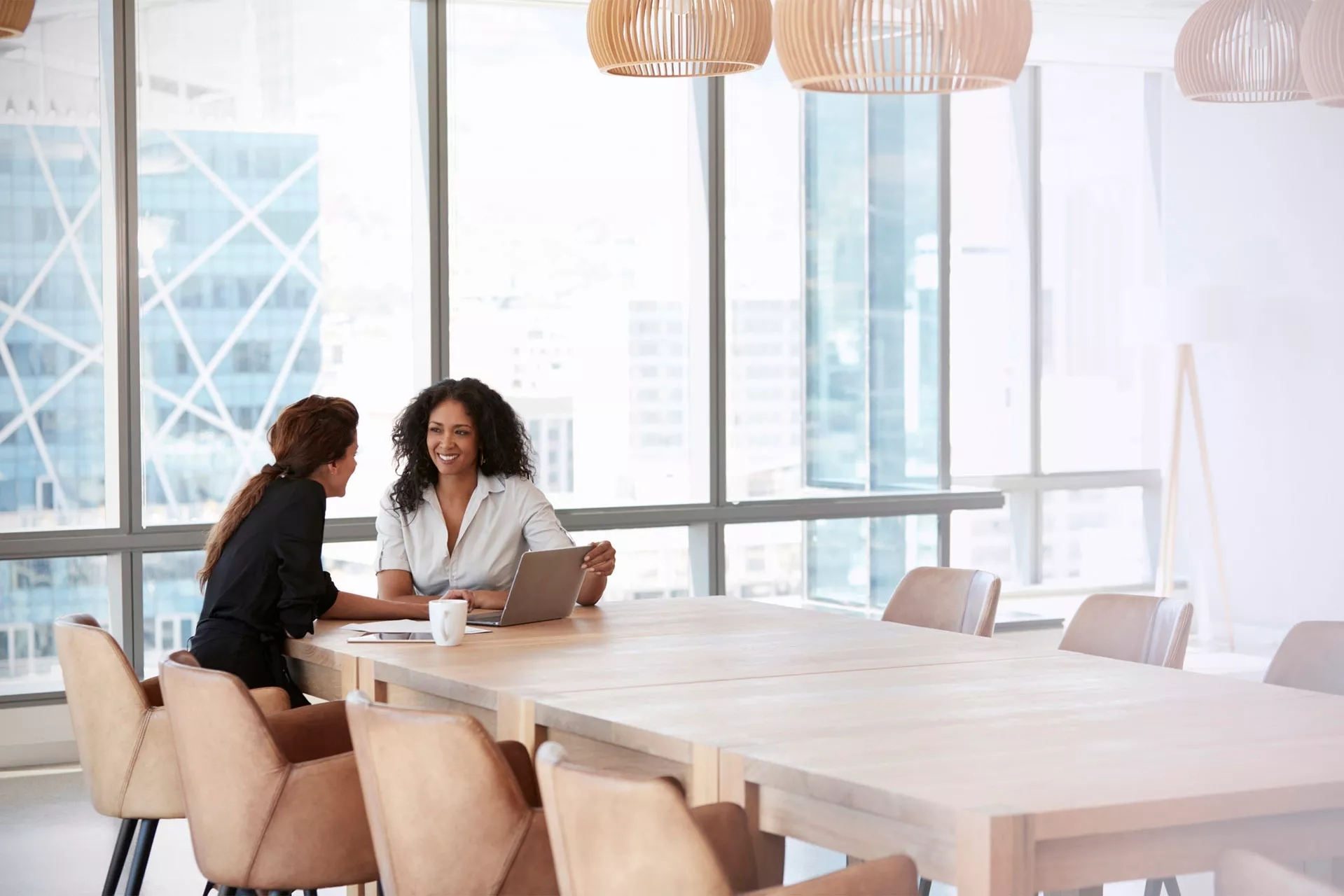 Two women meeting in a window-lined room, cityscape in the background. They are smiling, discussing the open laptop and notebook between them. Two women meeting in a window-lined room, cityscape in the background. They are smiling, discussing the open laptop and notebook between them.