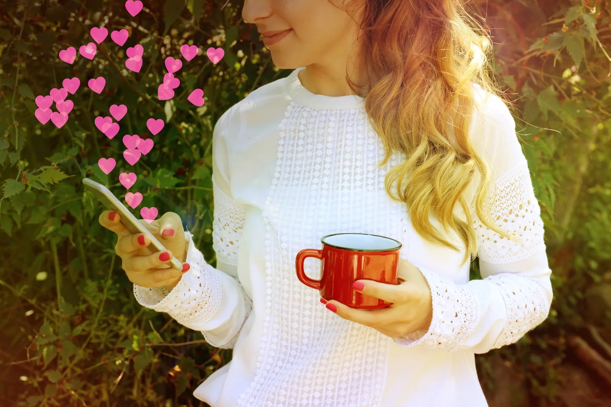 A woman is outside in front of some bushes, messaging on her phone, with heart emojis flying out, holding a mug in the other hand. A woman is outside in front of some bushes, messaging on her phone, with heart emojis flying out, holding a mug in the other hand.